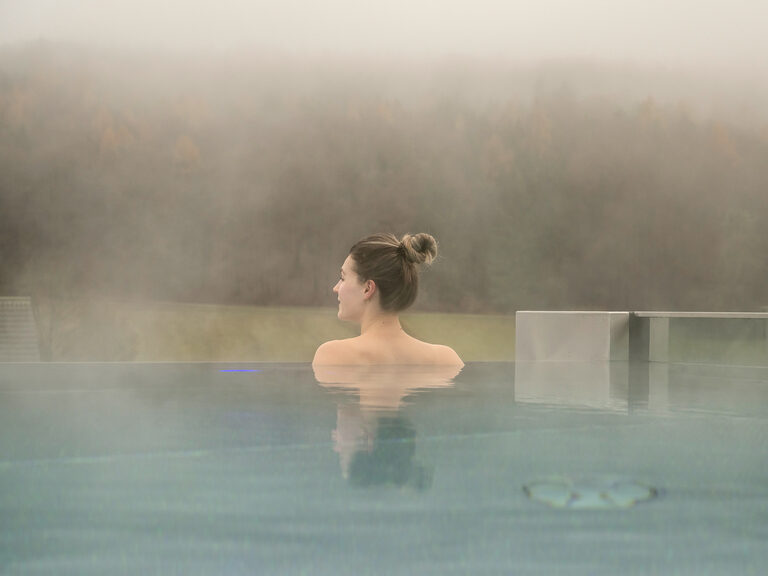 Eine Frau schaut über den Rand eines dampfenden Swimmingpools in die Ferne über die Wälder der Oberpfalz.