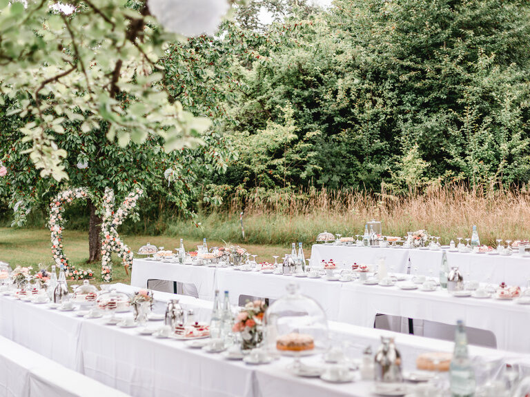 Lange schön dekorierte Hochzeitstafeln stehen auf einer Wiese umgeben von Bäumen und im Hintergrund sind die Initialen des Brautpaares mit Blumen geschmückt zu erkennen.