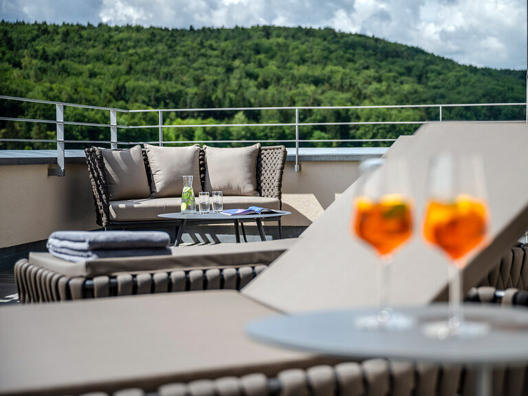 Zwei Cocktails stehen auf einem Tisch neben einer Sonnenliege mit Blick über die Umgebung